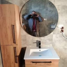 DF usługi remontowe Dawid Fyda - Nowoczesna łazienka z umywalką nablatową, drewnianą szafką pod umywalką i wysoką szafką boczną z drewnianymi frontami, okrągłe lustro z widocznym odbiciem fotografa robiącego zdjęcie.