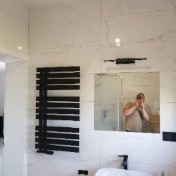 DF usługi remontowe Dawid Fyda - Wykończona łazienka z białymi marmurkowymi płytkami, czarnym grzejnikiem drabinkowym, owalną białą umywalką nablatową z czarną baterią i lustrem, w którym odbija się osoba robiąca zdjęcie.