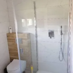 Nowoczesna kabina prysznicowa z deszczownicą i panelem prysznicowym, obok toaleta z zabudową drewnopodobną i białe płytki imitujące marmur na ścianach.