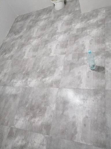 Świeżo położone, szare płytki podłogowe imitujące beton, z widocznym wiadrem po farbie i butelką wody, ukazujące etap prac wykończeniowych w pomieszczeniu.