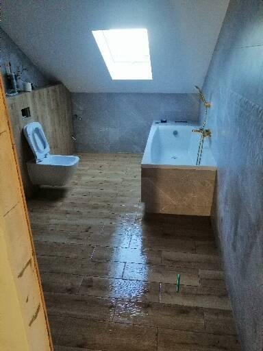 Ukończona łazienka na poddaszu z drewnopodobnymi płytkami na podłodze, białą wanną i toaletą. Na podłodze widoczne ślady wilgoci.