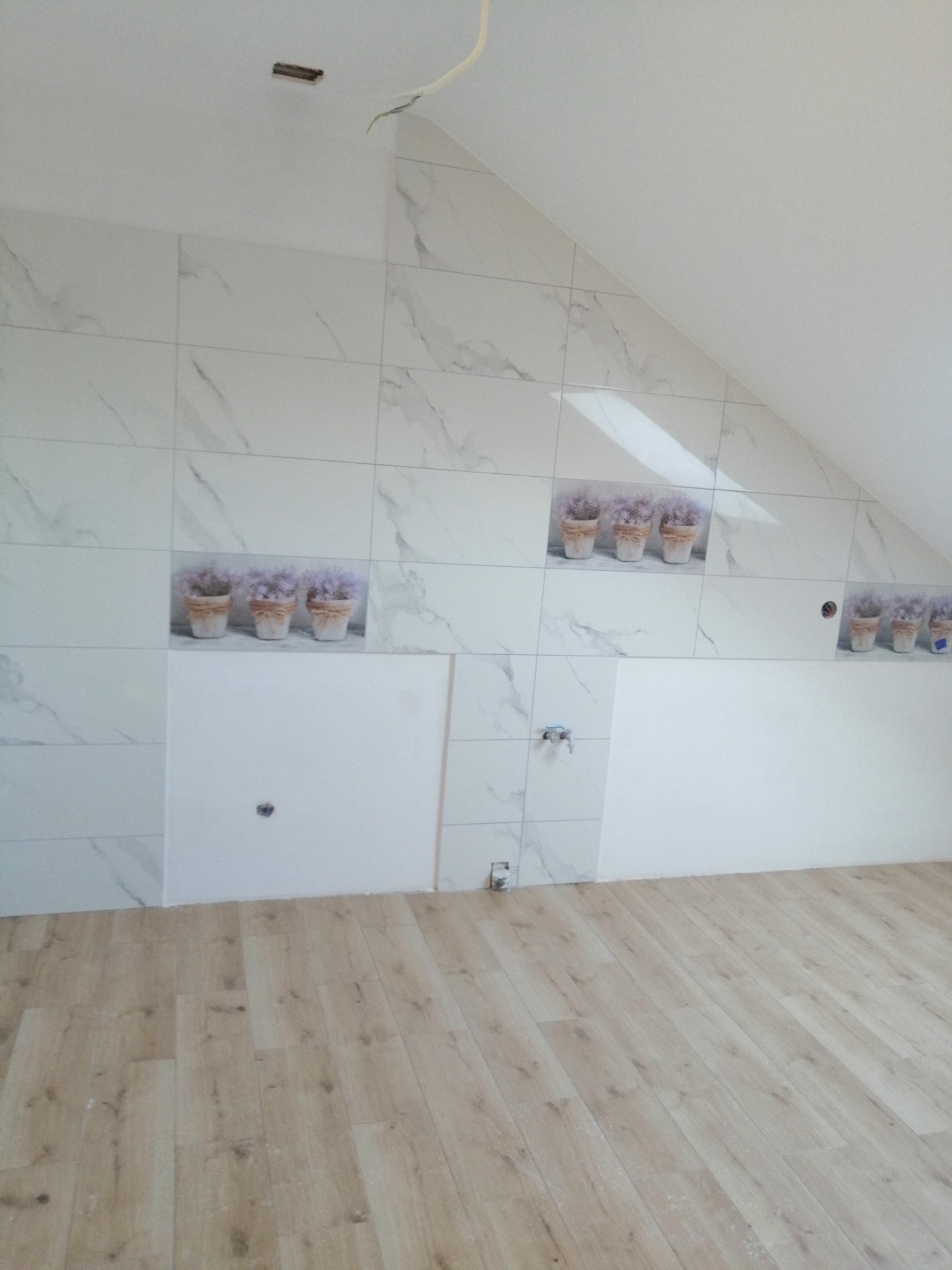 Ściana w łazience na poddaszu, wyłożona białymi płytkami imitującymi marmur z dekoracyjnymi wstawkami z motywem lawendy w doniczkach, częściowo jeszcze nieukończona, z widocznymi otworami...