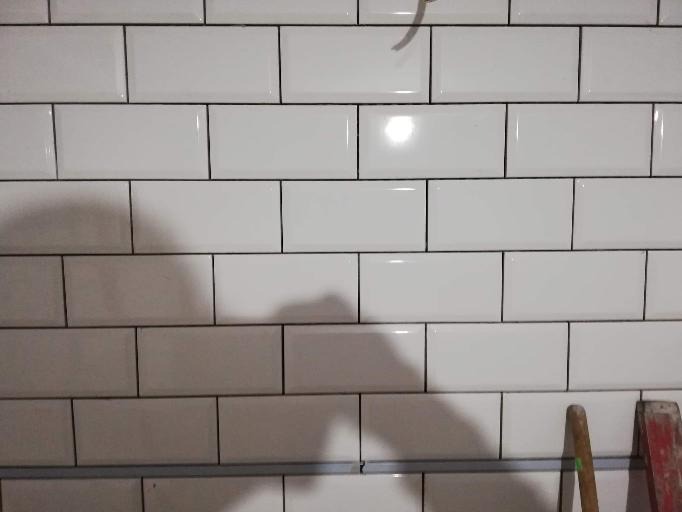 Ściana wyłożona białymi, prostokątnymi płytkami z ciemną fugą. W dolnej części widoczny fragment drabiny i listwa poziomująca.