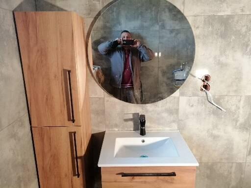 Nowoczesna łazienka z umywalką nablatową, drewnianą szafką pod umywalką i wysoką szafką boczną z drewnianymi frontami, okrągłe lustro z widocznym odbiciem fotografa robiącego zdjęcie.