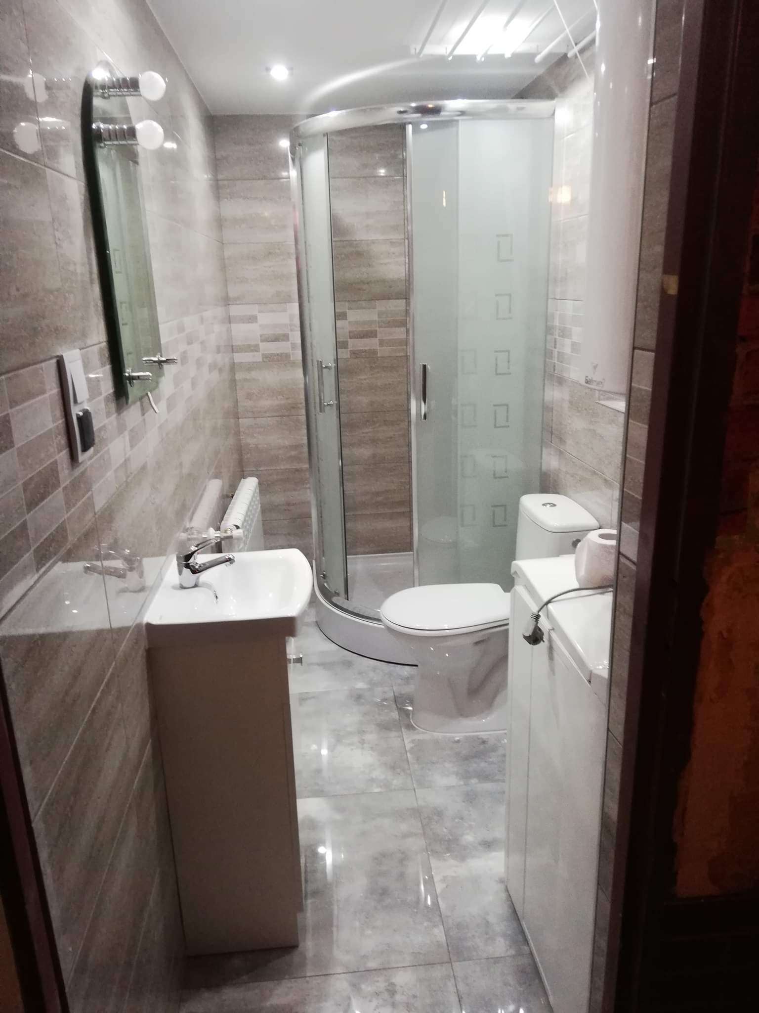 Nowoczesna łazienka z beżowymi płytkami, kabiną prysznicową z mlecznego szkła i umywalką na szafce, widziana zza framugi.