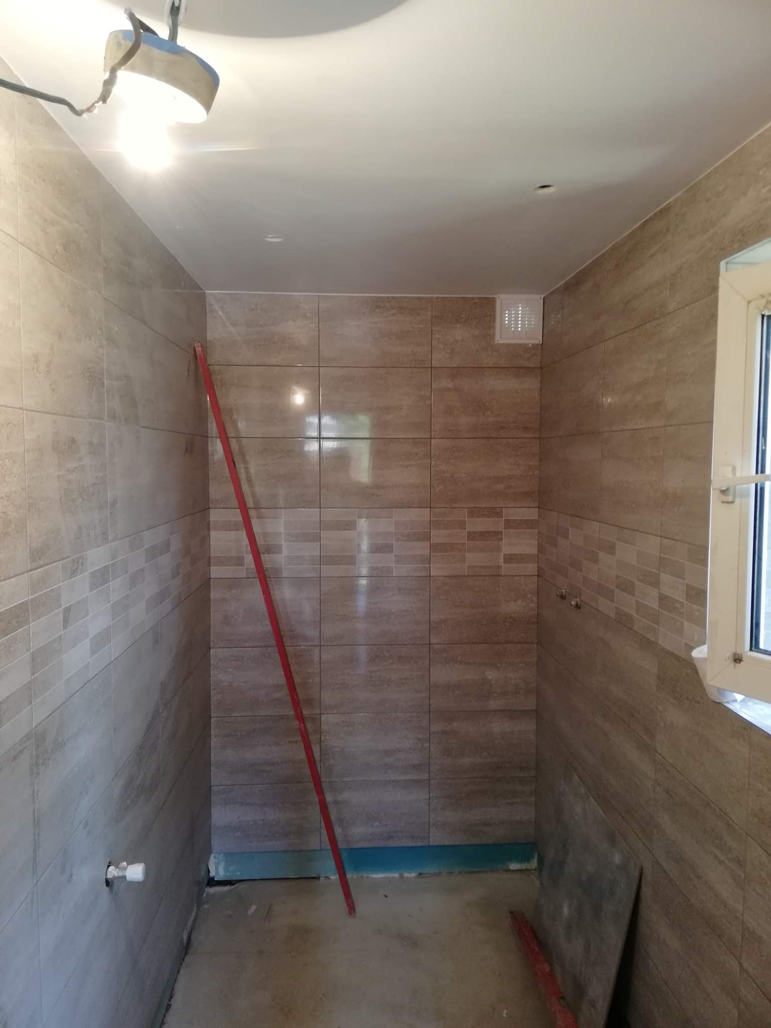 Świeżo wykafelkowana łazienka z poziomymi płytkami w odcieniach beżu i brązu, zabezpieczona folią w dolnej części ścian, z prowizorycznym oświetleniem budowlanym i poziomnicą opartą o ścianę.