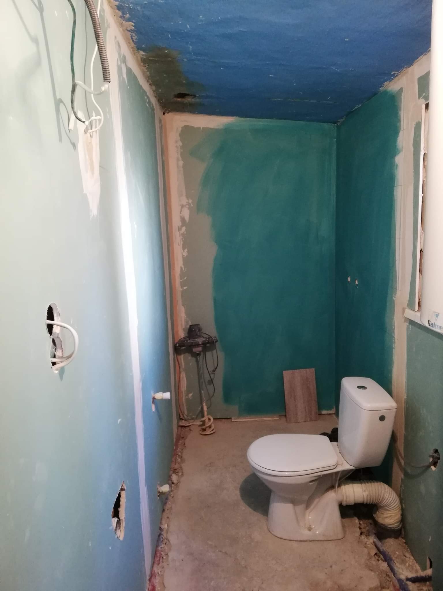 Łazienka w trakcie remontu: pomalowane na zielono ściany, niebieski sufit, zamontowana toaleta, widoczne kable i otwory w ścianach, mieszadło do farby na podłodze.