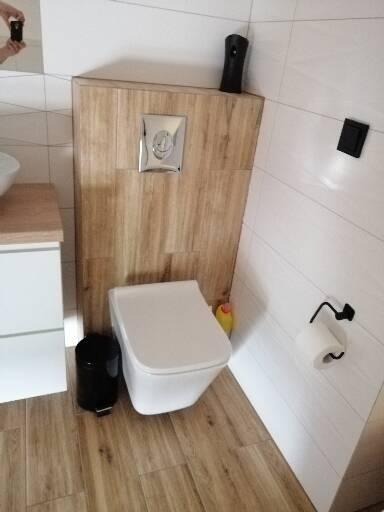 Nowoczesna toaleta w łazience z płytkami imitującymi drewno, zabudowany stelaż WC z chromowanym przyciskiem spłukiwania, czarny kosz na śmieci i pojemnik z odświeżaczem powietrza.