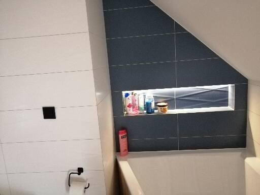 Wykończona łazienka z ciemnymi płytkami na ścianie pod skosem, wnęka z oświetleniem LED i kosmetykami, białe płytki z czarnym włącznikiem światła i uchwytem na papier toaletowy.