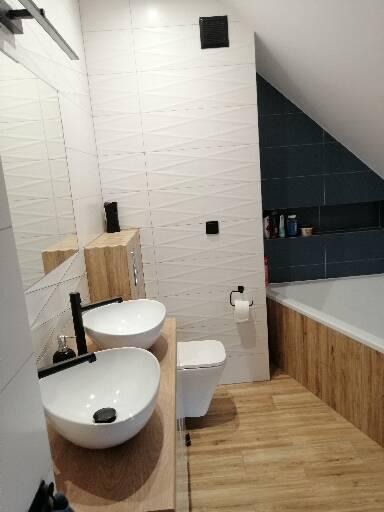 Nowoczesna łazienka na poddaszu z dwoma okrągłymi umywalkami na drewnianym blacie, białe płytki z subtelnym wzorem na ścianach i drewnopodobna okładzina wanny.