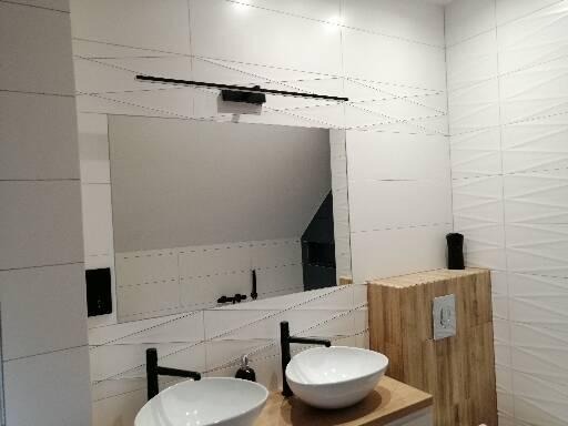 Nowoczesna łazienka z umywalkami nablatowymi, czarnymi bateriami i lustrem z oświetleniem LED, wykończona białymi płytkami z geometrycznym wzorem 3D na ścianie.