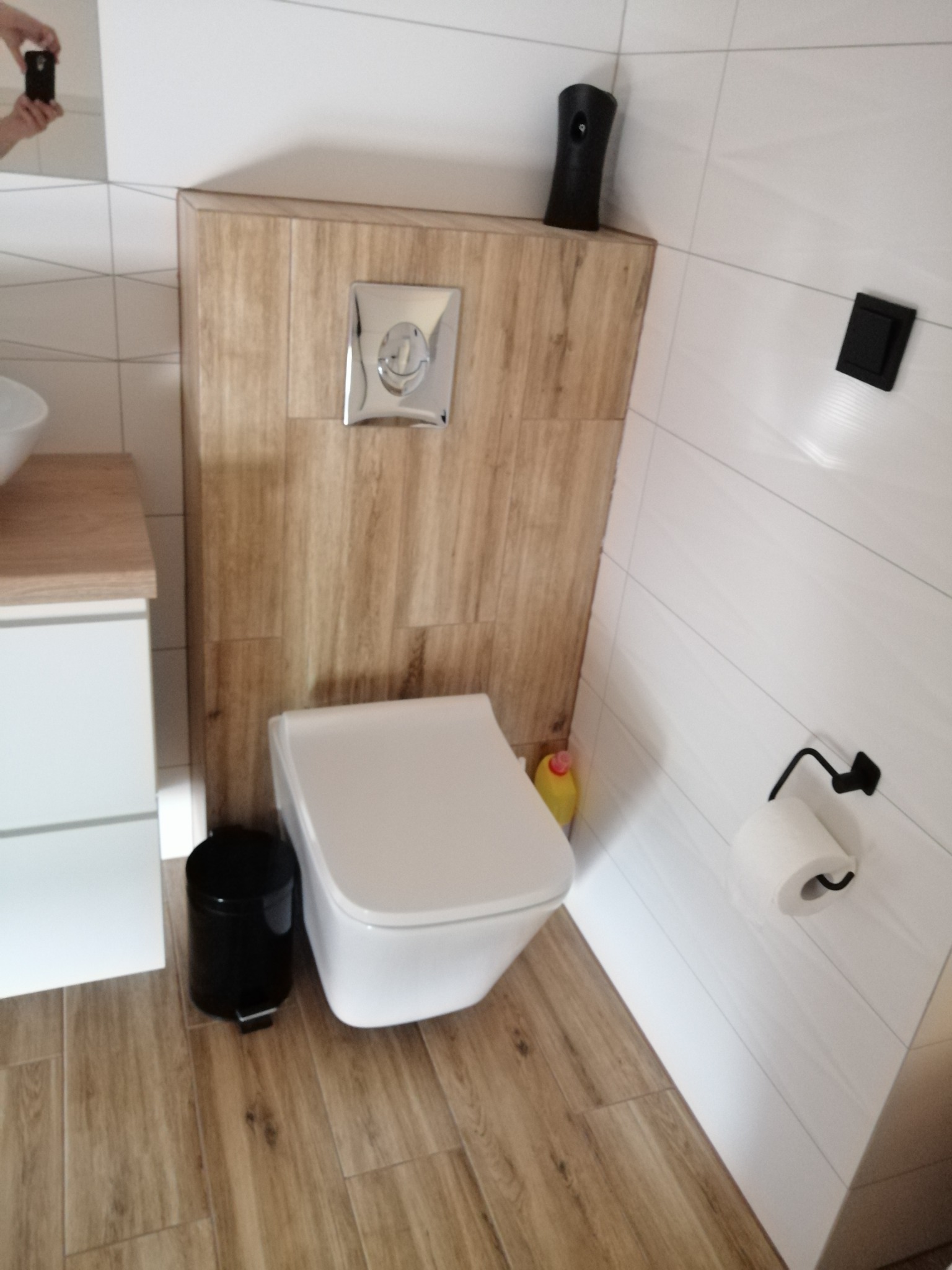 Nowoczesna toaleta podwieszana z drewnopodobną obudową stelaża, chromowany przycisk spłukujący, czarny pojemnik na śmieci i uchwyt na papier toaletowy w minimalistycznej łazience z białymi płytkami.