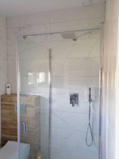 Kabina prysznicowa z deszczownicą i baterią podtynkową, wykończona białymi płytkami imitującymi marmur, z elementami zabudowy z płytek drewnopodobnych.