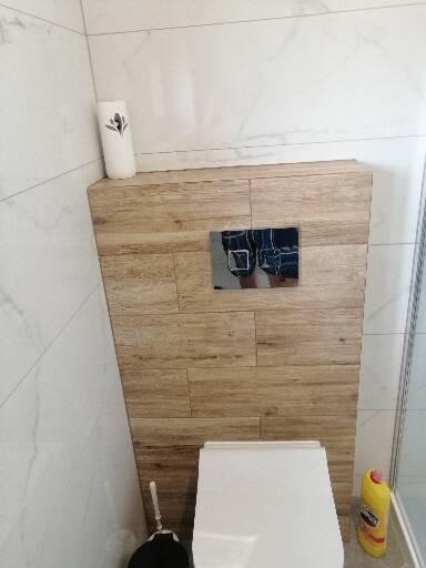 Świeżo wykończona toaleta podwieszana z obudową z płytek imitujących drewno, przycisk spłukujący w kolorze chromu, widoczna szczotka do WC i butelka płynu czyszczącego w rogu.