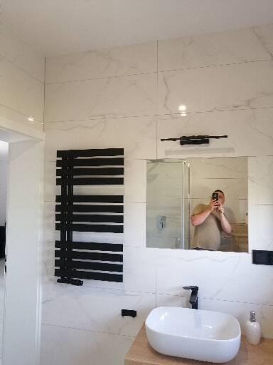 Wykończona łazienka z białymi marmurkowymi płytkami, czarnym grzejnikiem drabinkowym, owalną białą umywalką nablatową z czarną baterią i lustrem, w którym odbija się osoba robiąca zdjęcie.