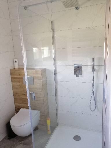 Nowoczesna kabina prysznicowa z deszczownicą i panelem prysznicowym, obok toaleta z zabudową drewnopodobną i białe płytki imitujące marmur na ścianach.