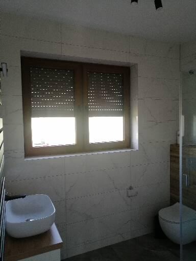 Wykończona łazienka z marmurowymi płytkami, umywalką nablatową i oknem z roletami zewnętrznymi.