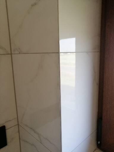 Ściana wyłożona białymi, marmurkowymi płytkami ceramicznymi, z widocznymi fugami i odbiciem światła na gładkiej powierzchni, przylegająca do drewnianej framugi drzwi.