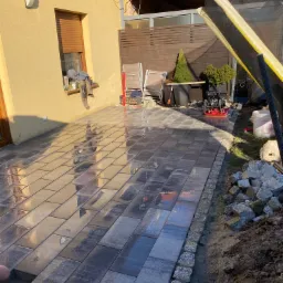 Układanie szarej prostokątnej kostki na tarasie, widoczne narzędzia i obrzeża z kostki granitowej, częściowo ułożona powierzchnia odbija światło słoneczne.