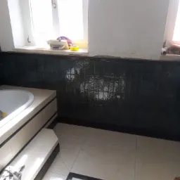 Fragment łazienki w trakcie prac: ułożone ciemnozielone, błyszczące płytki na ścianie nad wanną, jasna podłoga z ciemnymi akcentami, widoczne narzędzia i wiaderko z zaprawą, ślady kleju na ścianie,...