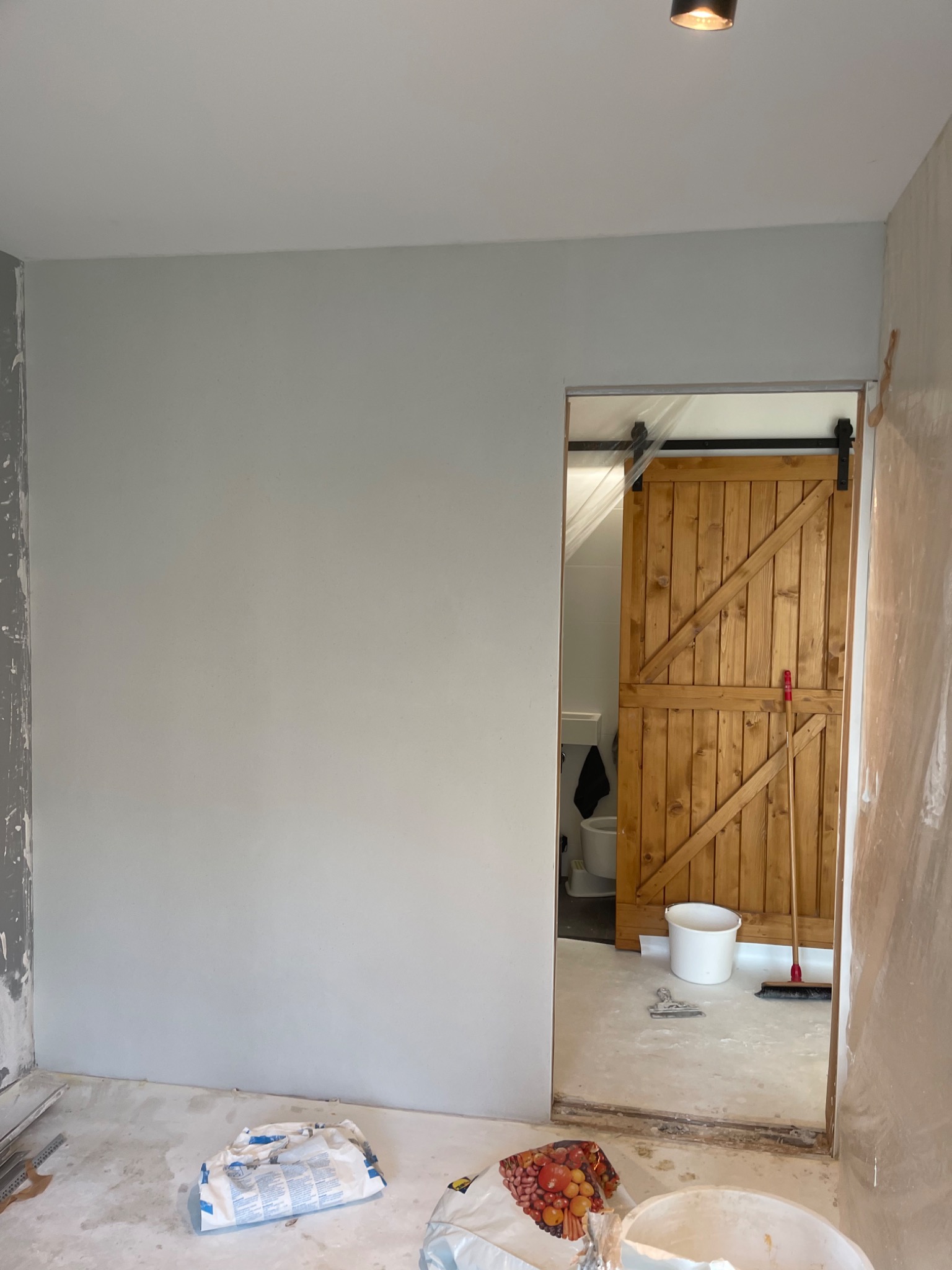 Pokój w trakcie remontu z przygotowaną, szarą ścianą do malowania, widoczne worki z zaprawą na podłodze oraz drewniane drzwi przesuwne do łazienki.