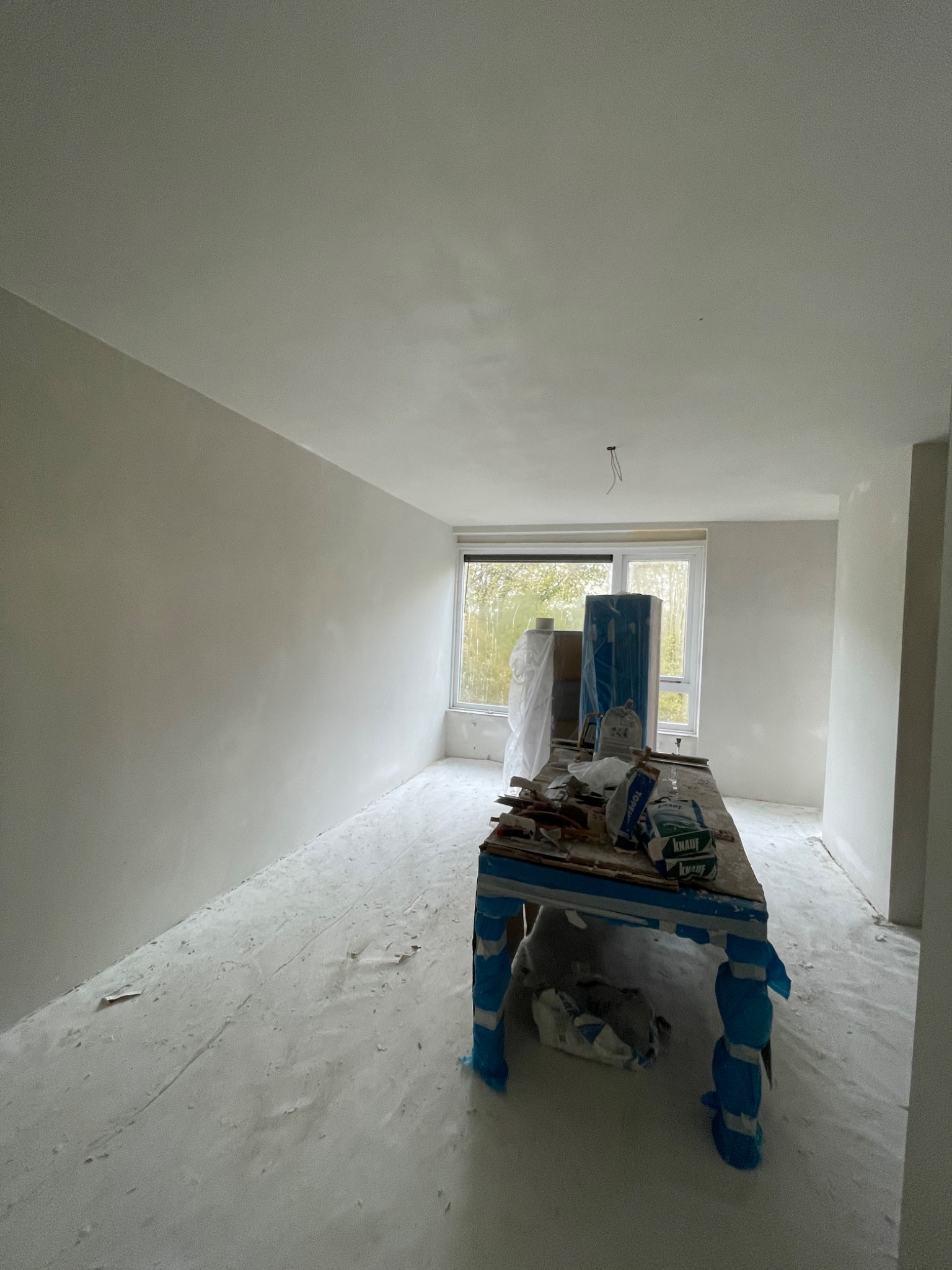 Pokój po nałożeniu gładzi, z widocznym stołem roboczym zabezpieczonym folią, workami z gładzią Knauf i narzędziami, podłoga przykryta warstwą pyłu, surowe oświetlenie z okna.