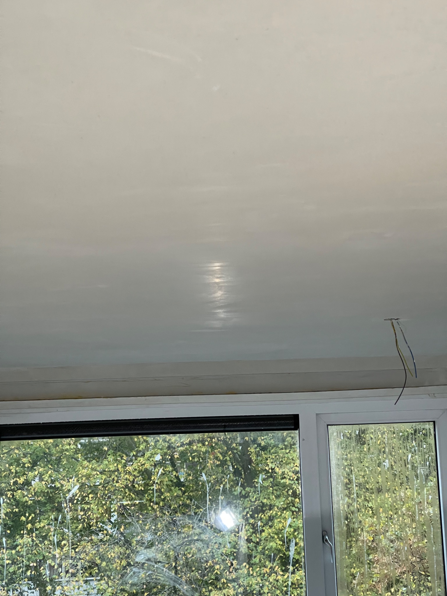 Gładki, kremowy sufit po szpachlowaniu, widoczne refleksy światła, wystające kable instalacji elektrycznej, okno z widokiem na zieleń na dole kadru.