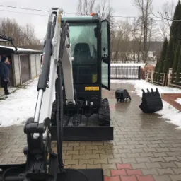 Kompaktowa minikoparka Bobcat z otwartą kabiną na kostce brukowej w pochmurny dzień; dwa dodatkowe łyżki obok, kobieta w kurtce w tle.