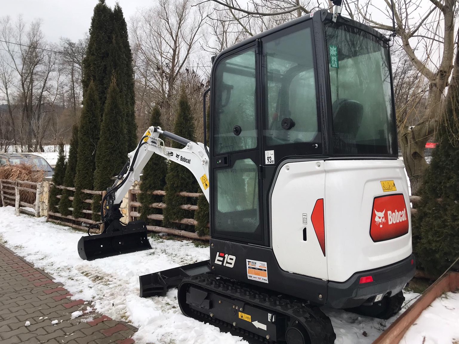 Minikoparka Bobcat E19 na śniegu, łyżka skarpowa zamontowana, widoczne ślady gąsienic na ziemi, w tle tuje i drewniany płot.