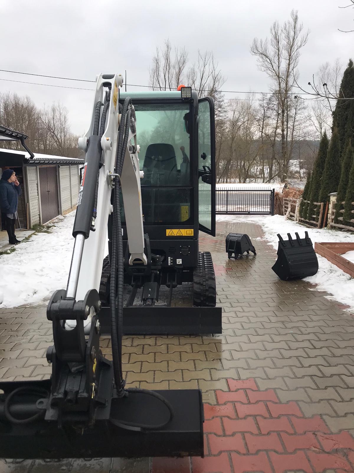 Kompaktowa minikoparka Bobcat z otwartą kabiną na kostce brukowej w pochmurny dzień; dwa dodatkowe łyżki obok, kobieta w kurtce w tle.