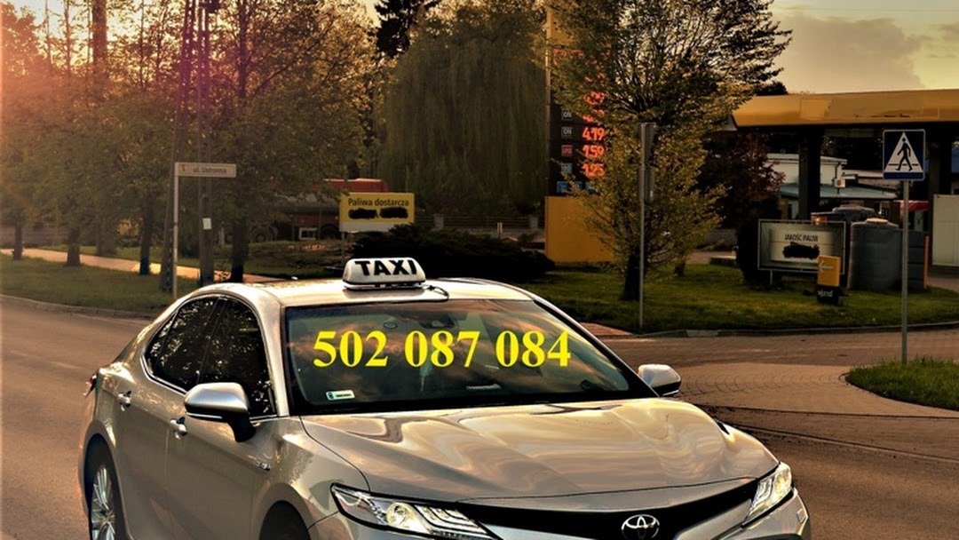 Srebrna taksówka Toyota z numerem telefonu na przedniej szybie, zaparkowana w pobliżu stacji benzynowej w słoneczny dzień.