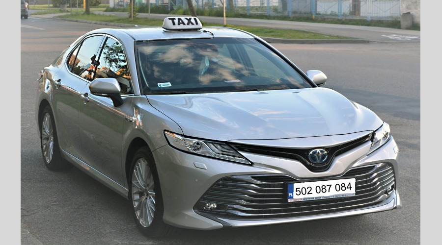 Srebrny samochód osobowy z napisem TAXI na dachu, zaparkowany na ulicy z widocznym numerem rejestracyjnym.