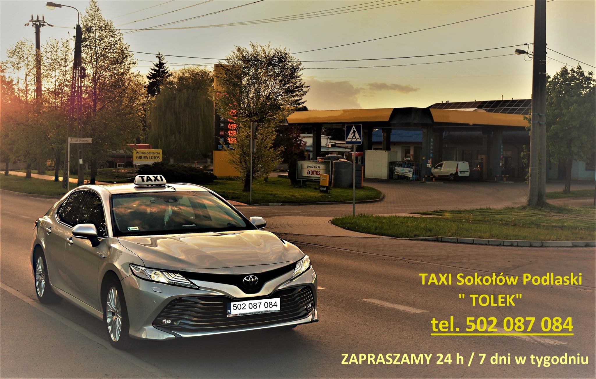 Srebrny samochód osobowy z napisem TAXI na dachu, zaparkowany na ulicy w Sokołowie Podlaskim, w tle stacja benzynowa LOTOS i drzewa w promieniach zachodzącego słońca.