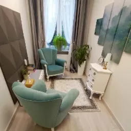 Ustawienie gabinetu z dwoma turkusowymi fotelami, białym stolikiem nocnym z lampką, obrazami górskimi, dywanem i roślinami doniczkowymi przy oknie z zasłonami.