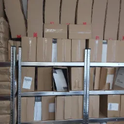 Półki regału magazynowego wypełnione kartonami, niektóre z naklejonymi etykietami adresowymi i taśmą klejącą.