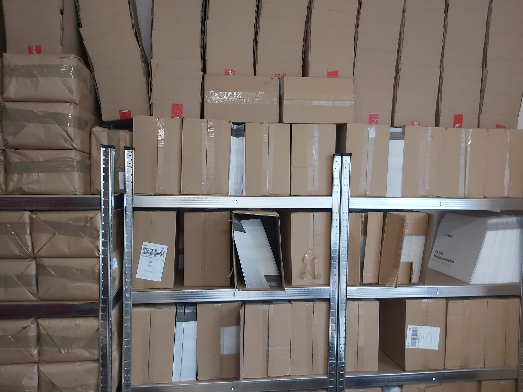 Półki regału magazynowego wypełnione kartonami, niektóre z naklejonymi etykietami adresowymi i taśmą klejącą.