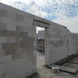 Wznoszenie ścian domu z betonu komórkowego, widoczne otwory okienne i drzwiowe, zbrojenie nadproży, w tle błękitne niebo z chmurami.