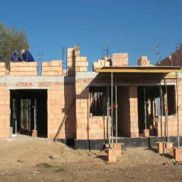 Budowa domu z cegły ceramicznej Porotherm, widoczni dwaj pracownicy na górze muru, podpory stropowe i żółta tablica informacyjna na ścianie budynku.