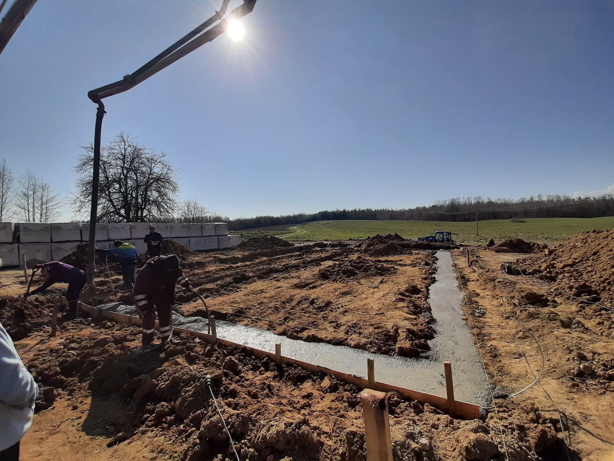 Wylewanie fundamentów z betonu na placu budowy, widoczny beton zbrojony w szalunku, pracownicy w odzieży roboczej, pompa do betonu, bloczki betonowe w tle i krajobraz polny.
