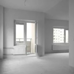 Pusty, jasny pokój w stanie deweloperskim z białymi ścianami, grzejnikami pod oknami i wyjściem na balkon, widok na budynek mieszkalny za oknem.