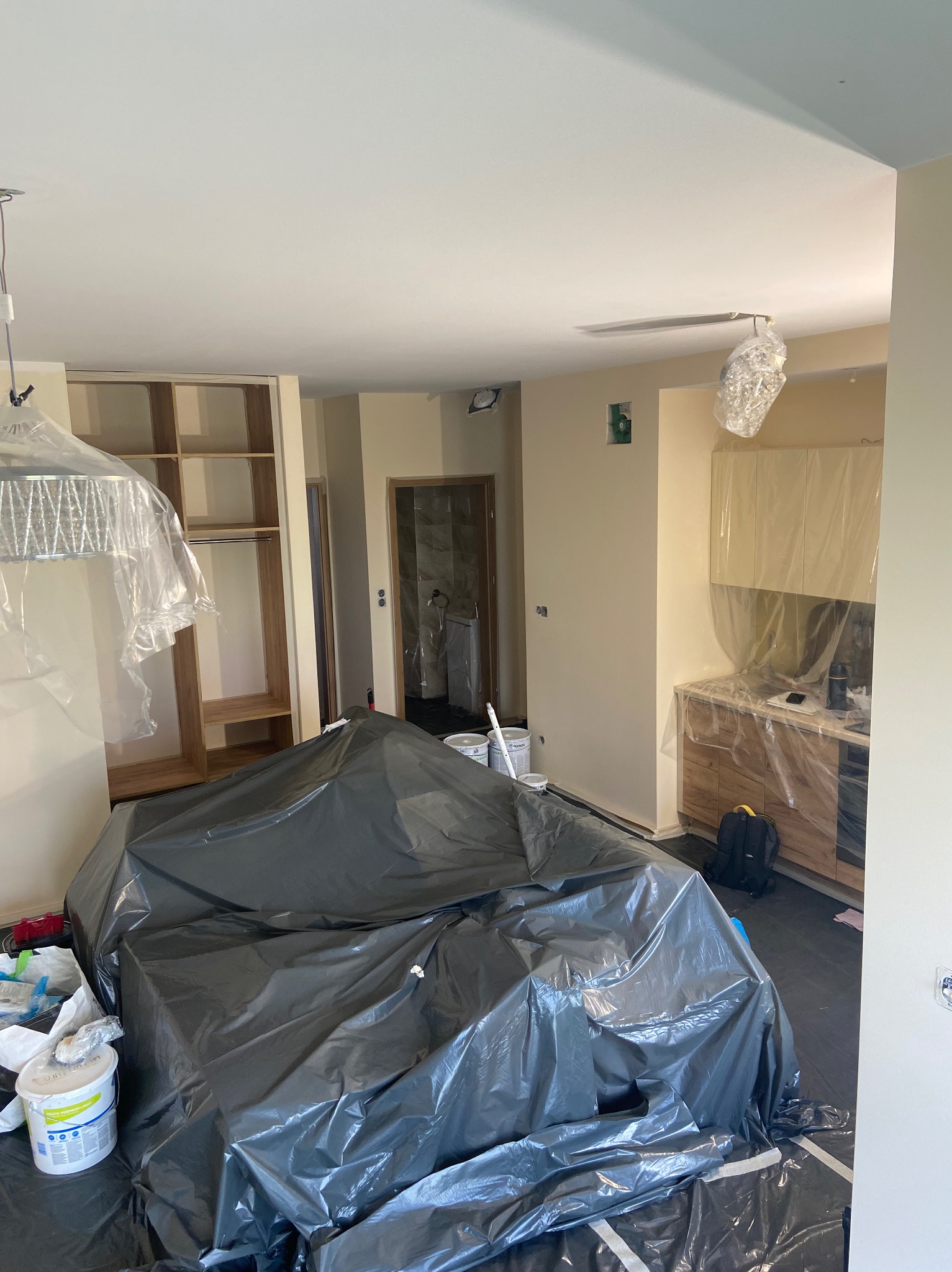 Pokój w trakcie przygotowań do prac wykończeniowych: meble zabezpieczone folią, ściany częściowo pomalowane na jasny kolor, widoczne puszki z farbą i akcesoria malarskie, podłoga zabezpieczona...