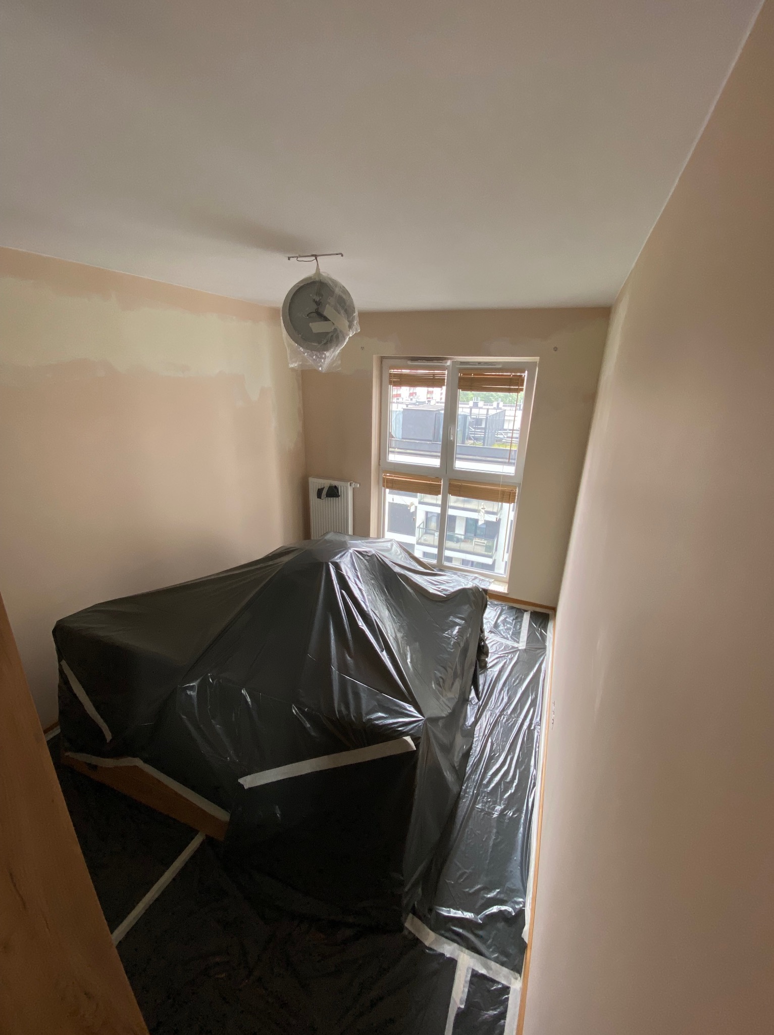 Pokój w trakcie przygotowań do prac wykończeniowych: meble zabezpieczone folią, ściany częściowo zagruntowane, zegar wiszący pod sufitem owinięty folią ochronną, okno z żaluzjami.