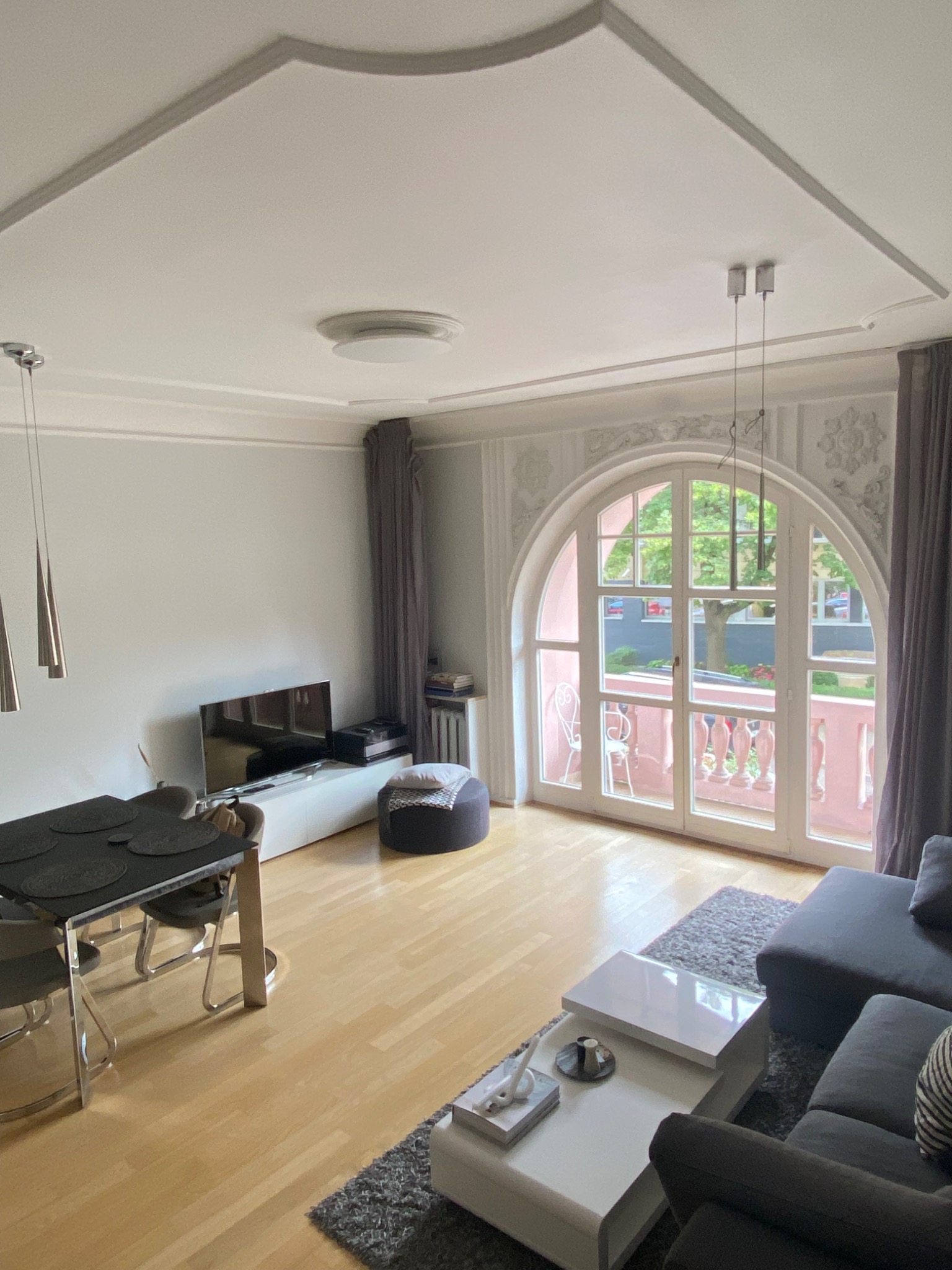 Jasne wnętrze salonu z dekoracyjnym sufitem i dużym, łukowym oknem balkonowym z różową balustradą, widok na zieleń za oknem, minimalistyczne meble.