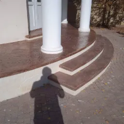 Krużganek z kolumnami i schodami wykończony wzorzystym, brązowym betonem stemplowanym, z widocznym cieniem osoby fotografującej.