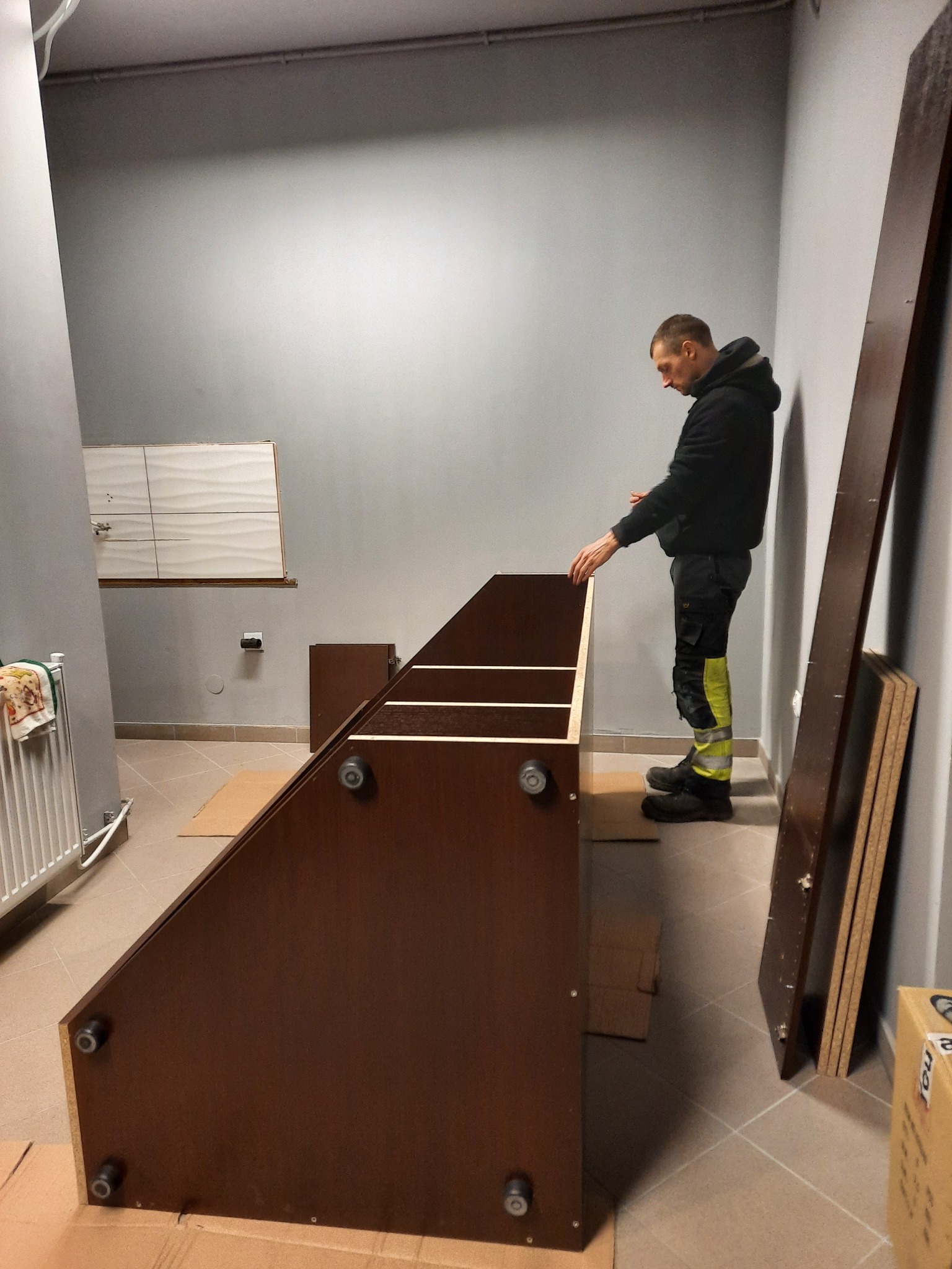 Mężczyzna w czarnej bluzie i spodniach roboczych montuje brązowy regał w szarym pomieszczeniu, widoczne elementy konstrukcyjne i kartonowe podkładki.