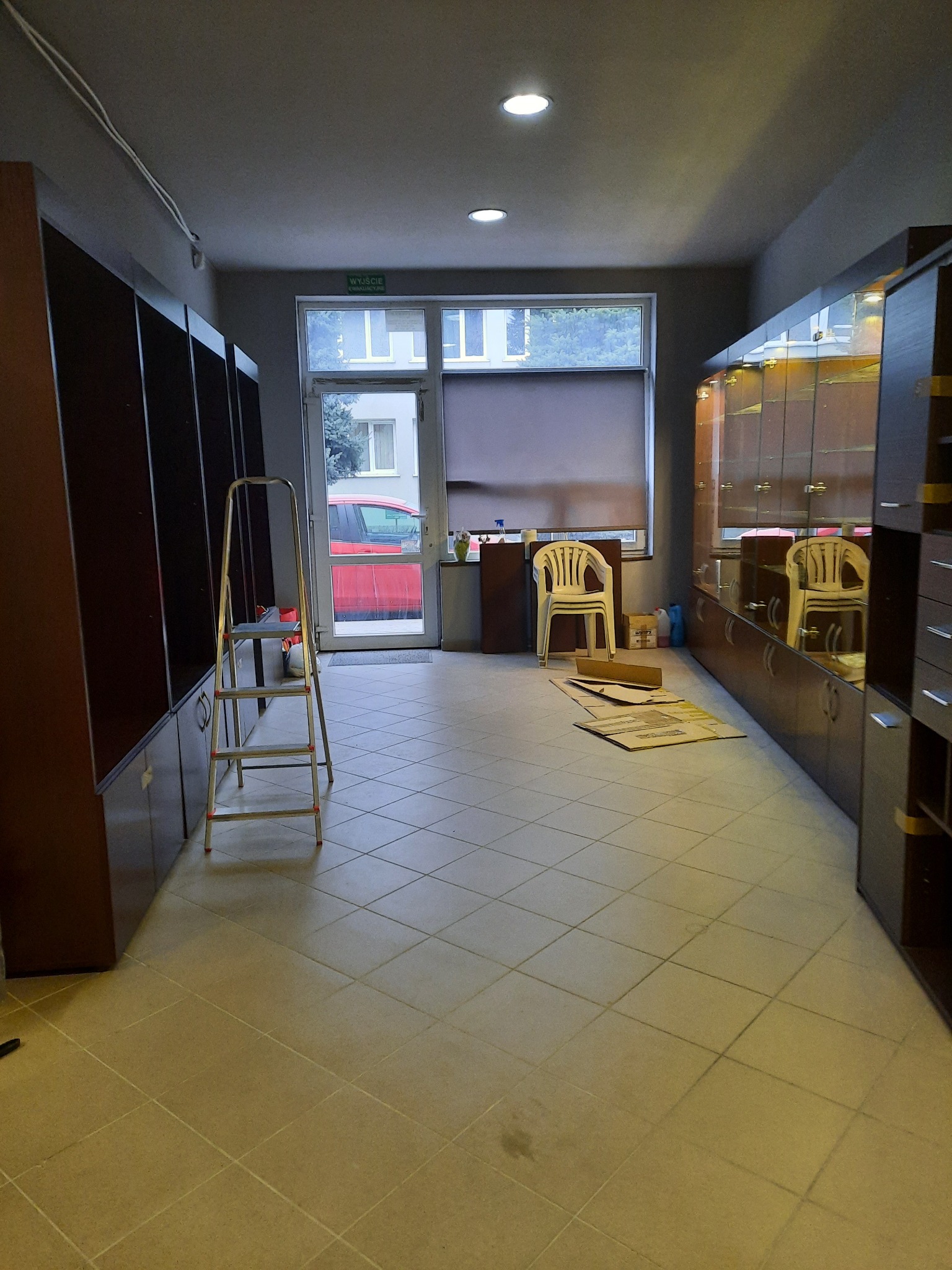 Wnętrze sklepu odzieżowego w trakcie remontu lub aranżacji, widoczne regały, witryny, drabina, kartony i krzesło.