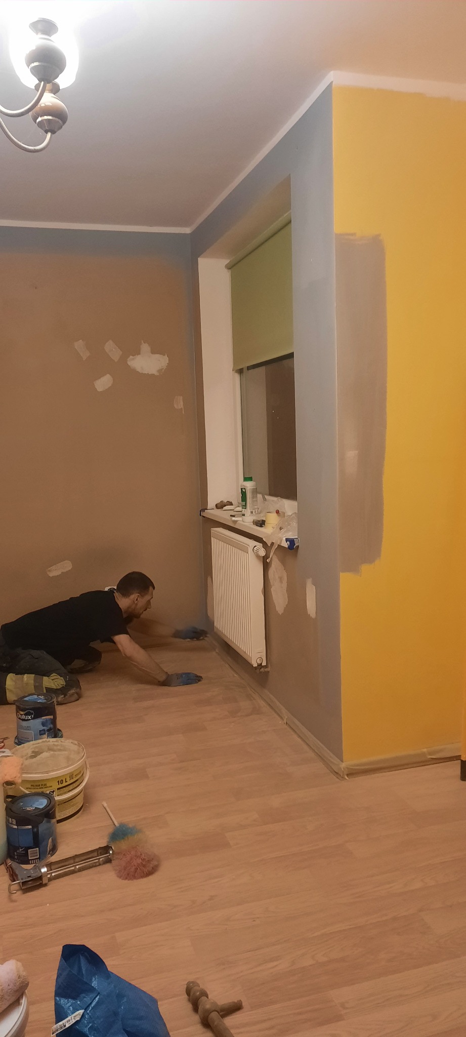 Mężczyzna w niebieskich rękawicach maluje dolną część ściany w pokoju na szaro, widoczne puszki z farbą, grzejnik i częściowo pomalowana na żółto ściana.