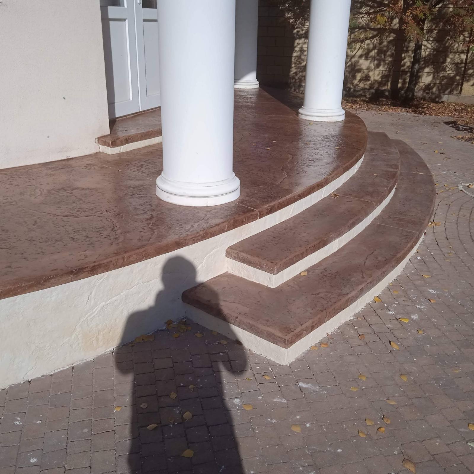 Krużganek z kolumnami i schodami wykończony wzorzystym, brązowym betonem stemplowanym, z widocznym cieniem osoby fotografującej.