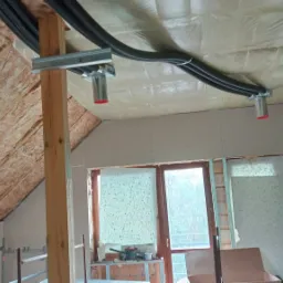 Wnętrze poddasza w trakcie montażu rekuperacji: widoczne czarne rury wentylacyjne, izolacja wełną mineralną, okno dachowe i częściowo zamontowane płyty gipsowo-kartonowe.