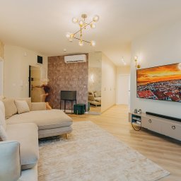 Zizi studio Magdalena Latos - Jasne, przestronne wnętrze salonu z beżową sofą narożną, wzorzystą tapetą, nowoczesnym oświetleniem i telewizorem z obrazem miasta o zachodzie słońca, osoba w ruchu w tle.